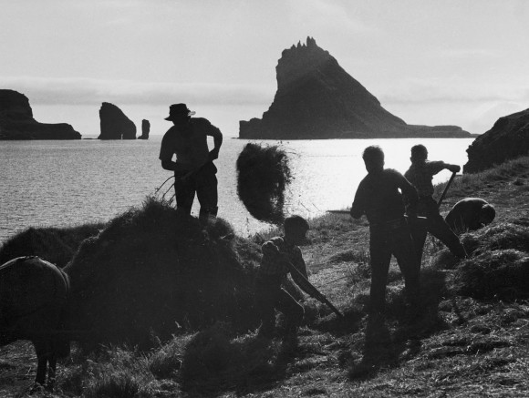 I “Høbjergning” (udateret) ses en færøsk bonde og hans tre unge hjælpere i silhuet mod det spejlblanke hav og den nedgående sol i horisonten. Foto: Ásmundur Poulsen.